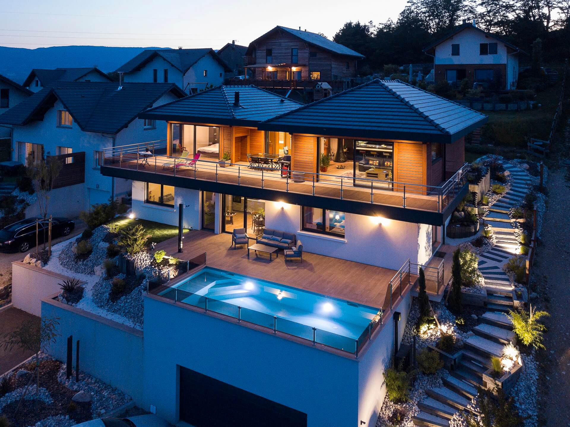 Vue nocturne d'une maison contemporaine en bois avec sous-sol en béton, grande terrasse, piscine éclairée et jardin paysager, située en Savoie.