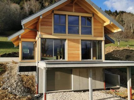 Maison contemporaine en bois avec de grandes ouvertures vitrées, toiture à deux pans et garage en sous-sol, construite en pente près du plateau des Petites Roches.