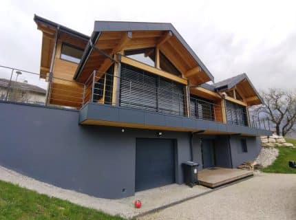Façade d’une maison contemporaine en bois avec habillages en aluminium, grandes ouvertures vitrées et garde-corps métalliques surélevés.
